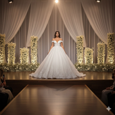 Model wearing a white wedding dress on a runway with floral decorations.