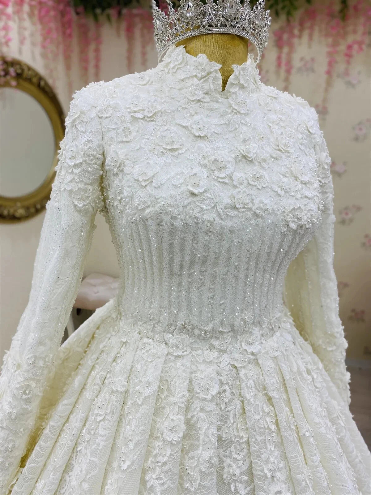 White lace wedding dress with long sleeves and a tiara on a mannequin against a floral wallpapered wall.